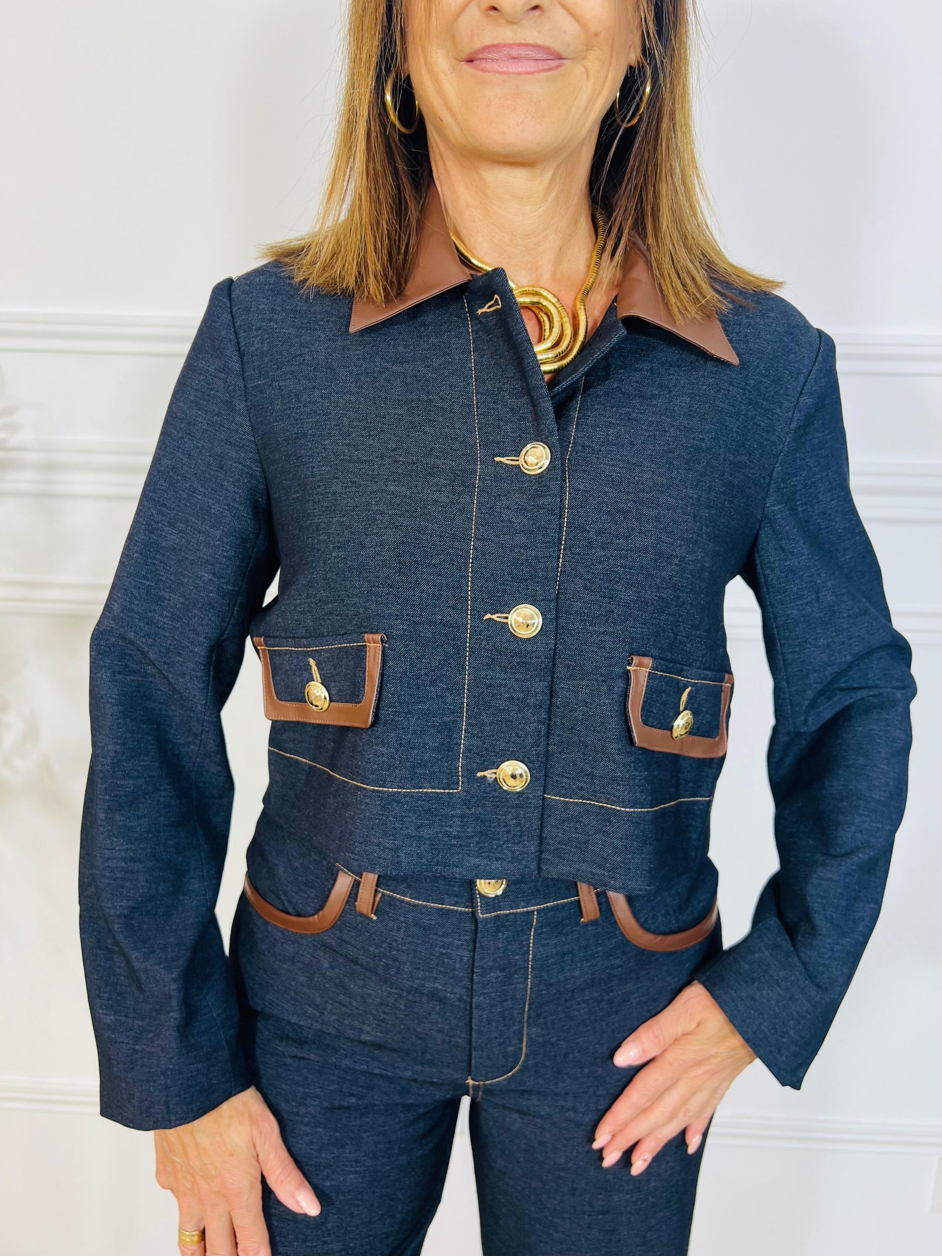 Chaqueta de corte recto y largo a la cintura, confeccionada en tejido tipo denim con un punto elástico que aporta comodidad y estilo. Presenta cuello camisero en polipiel marrón chocolate, bolsillos decorativos con solapa y botón dorado, y costuras en contraste que realzan el diseño. Cierre frontal con botones dorados de diseño elegante.